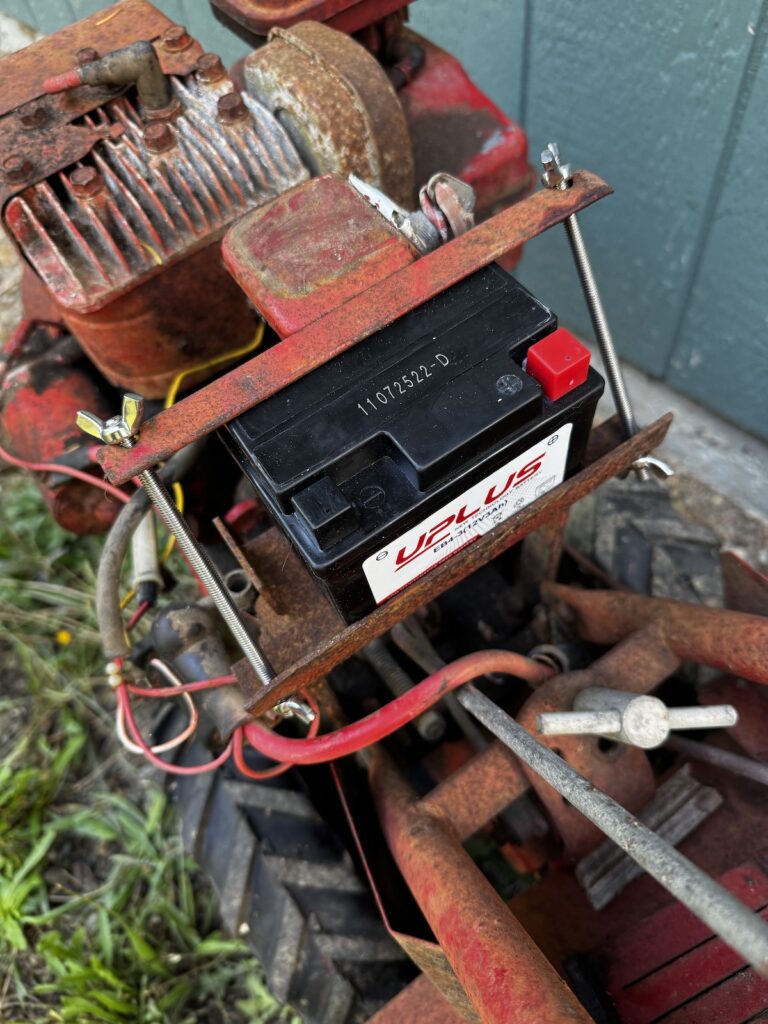 A red Troy-Built rototiller with a new battery held down by threaded rod and wing nuts with the original bracket.